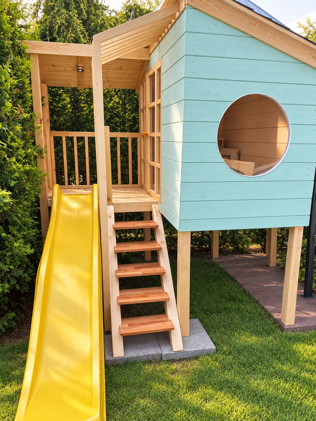 Daylight view of a raised mint-coloured playhouse with round window, yellow slide and timber steps